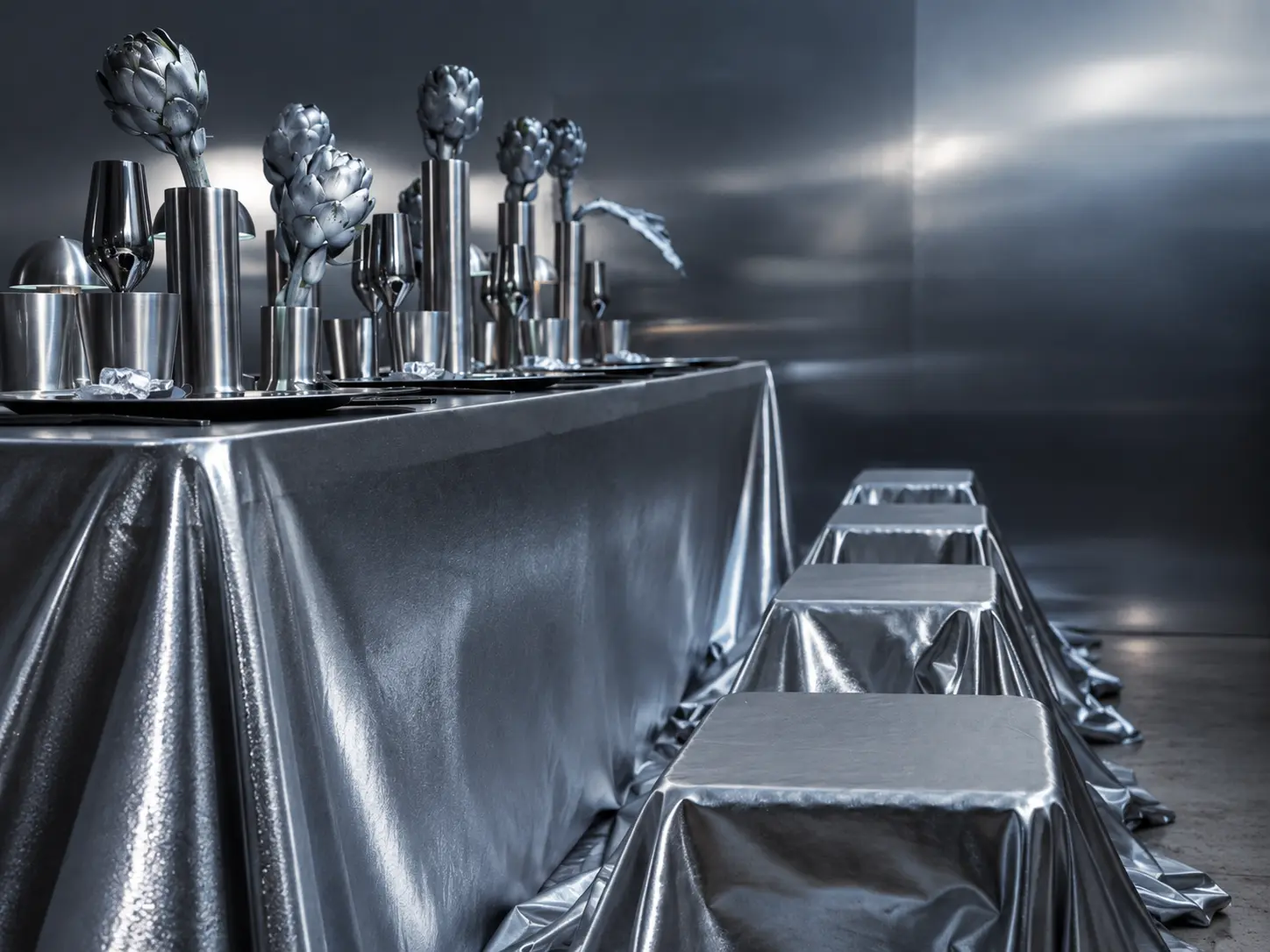 Contemporary event table with sculptural silver artichoke centerpieces, metallic cylindrical vases and silver-draped stools.