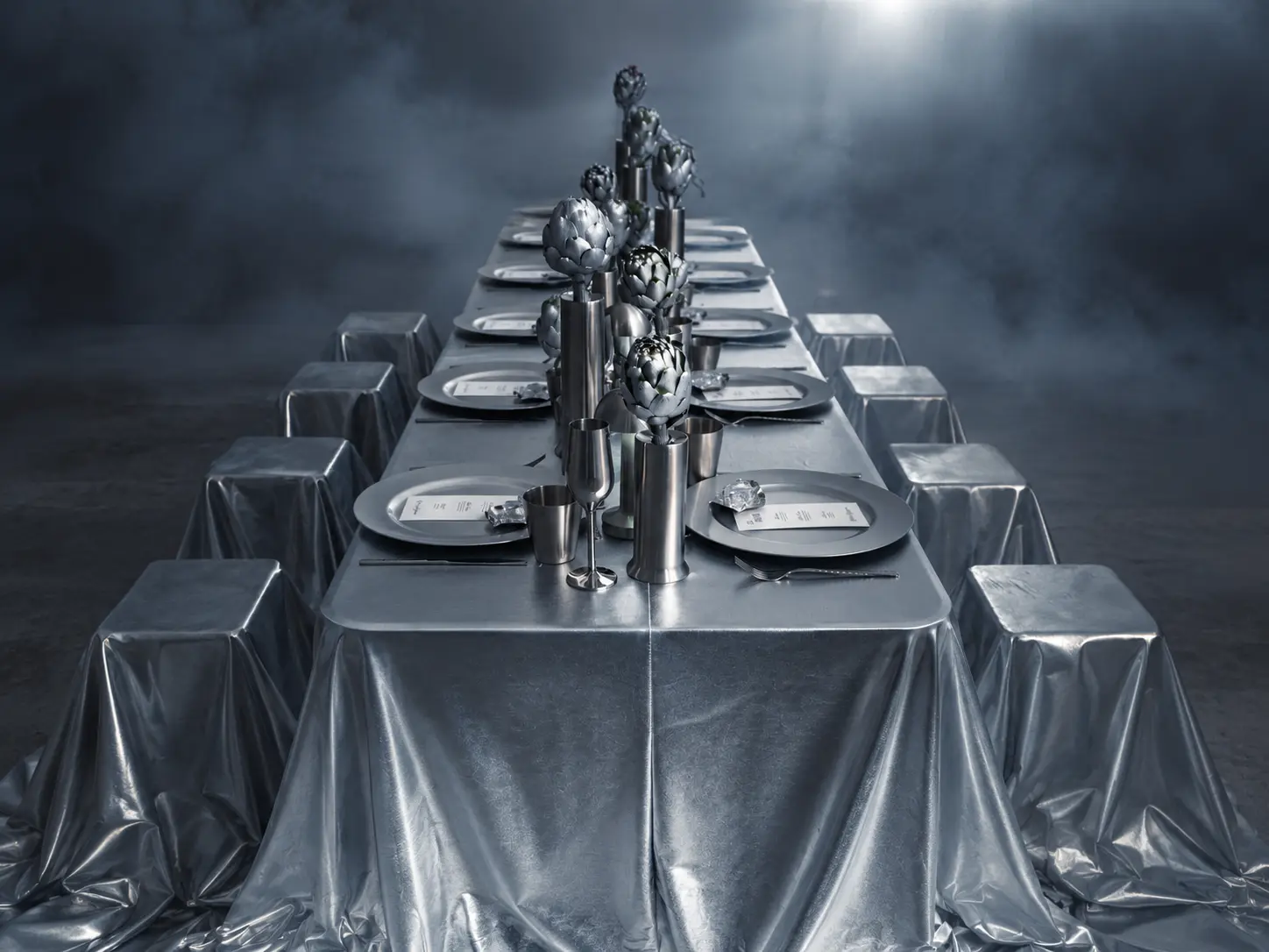 Symmetrical event table setting with metallic silver draping, sculptural artichoke centerpieces and silver-covered stools against a misty backdrop.