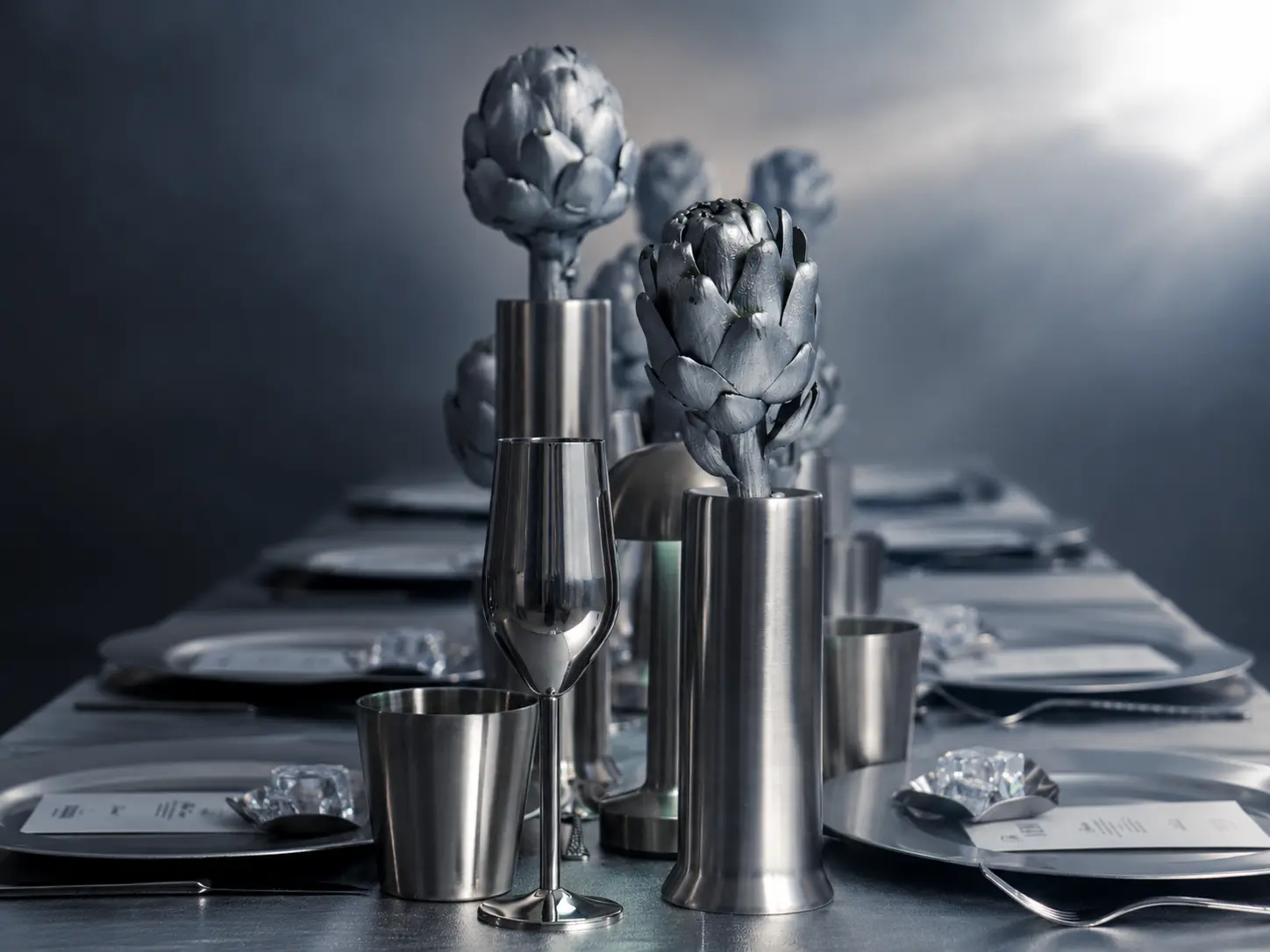 Close-up of silver artichoke centerpieces in metallic cylindrical vases, surrounded by modern silver tableware and glassware.