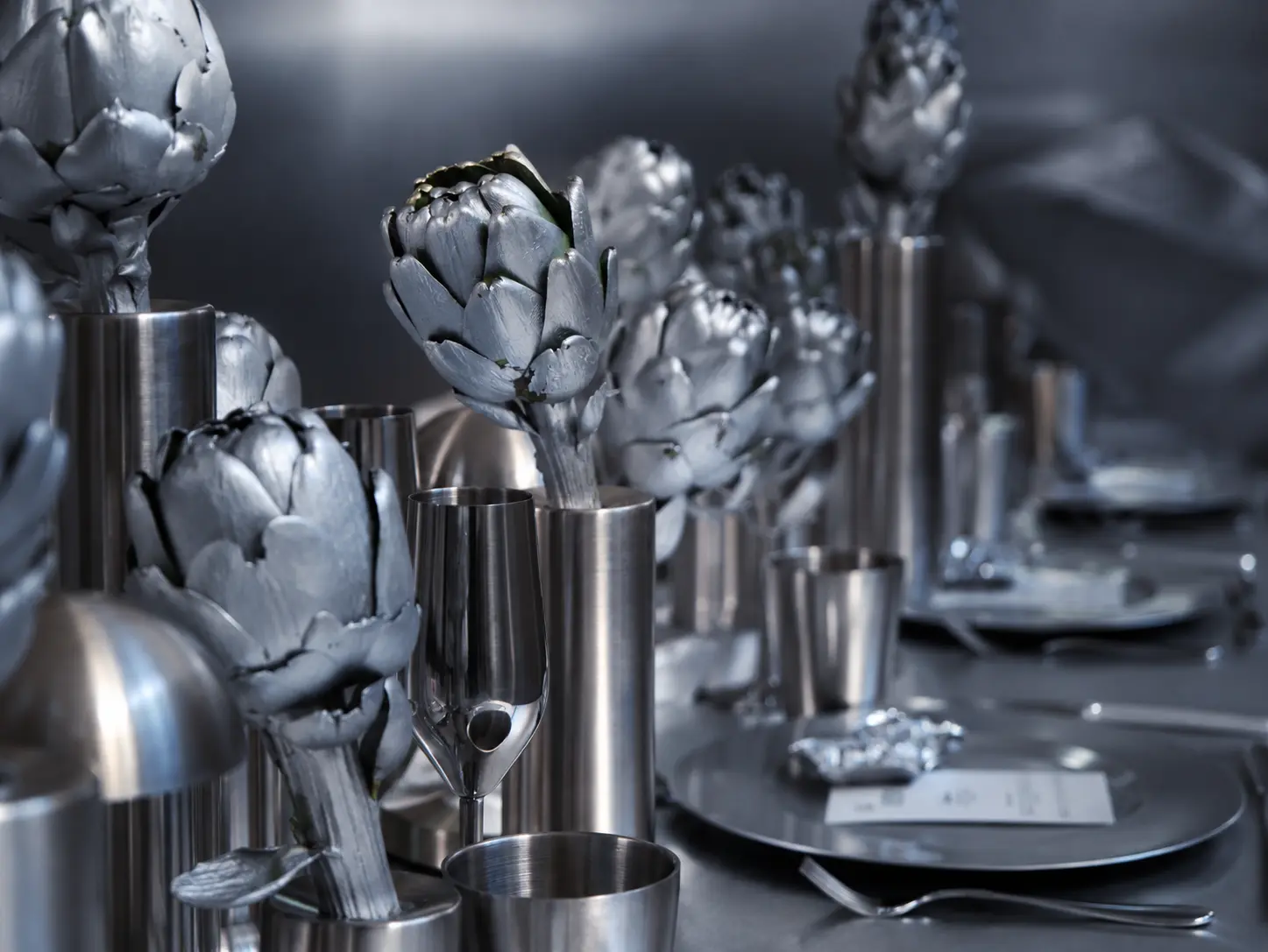 Close-up of sculptural silver artichokes arranged in metallic cylindrical vases, alongside silver glassware and contemporary table settings.