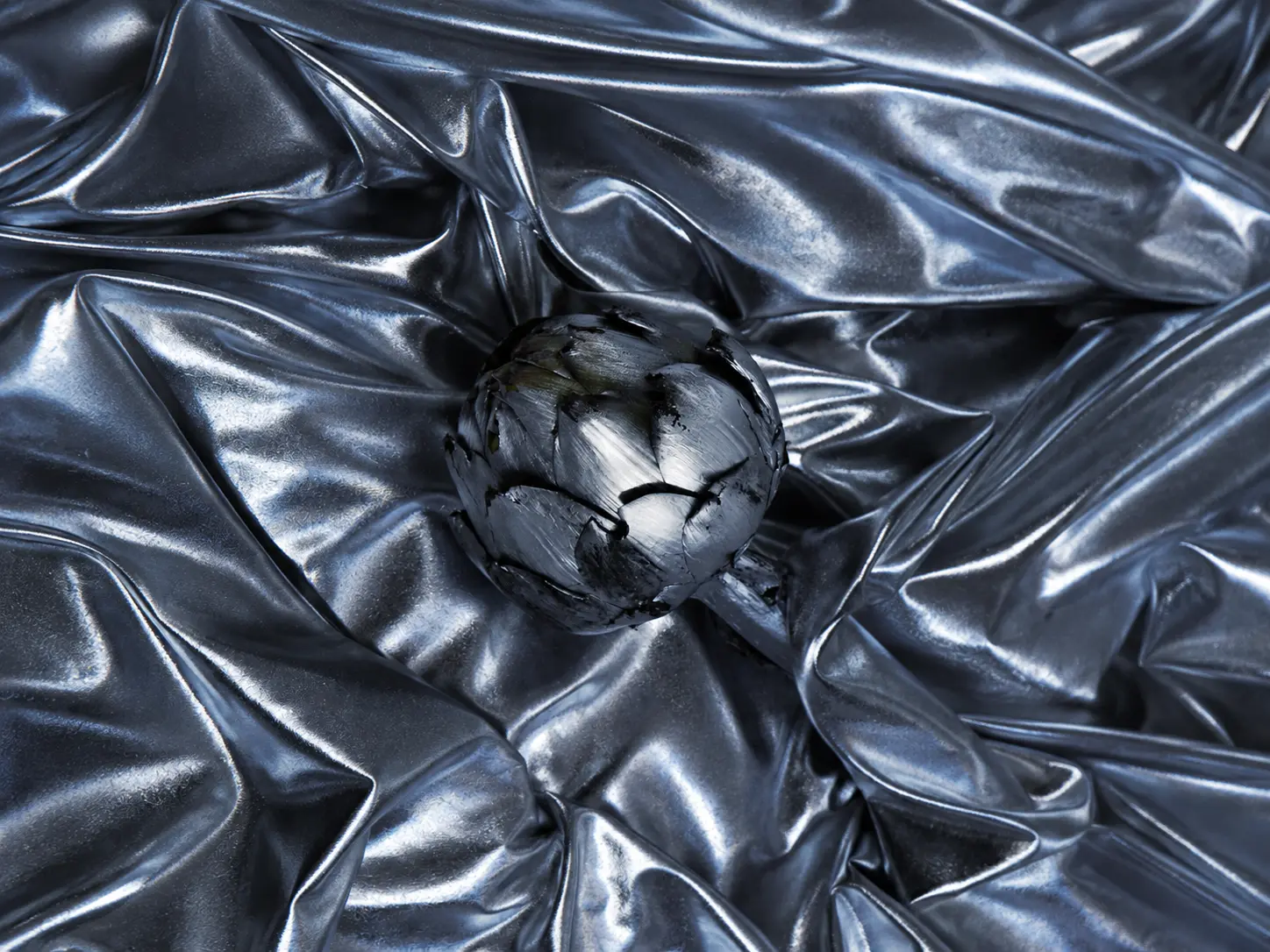 Single metallic silver artichoke surrounded by sculptural folds of reflective silver fabric in a monochromatic editorial composition.