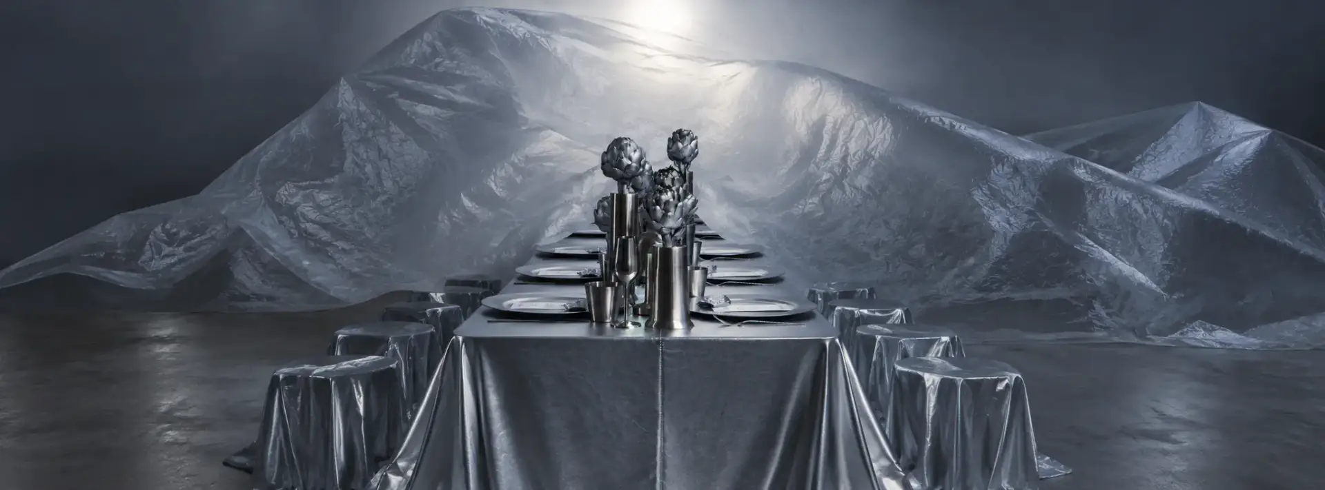 Futuristic event tablescape with metallic silver draping, sculptural artichoke centerpieces and geometric silver stools against an illuminated backdrop.