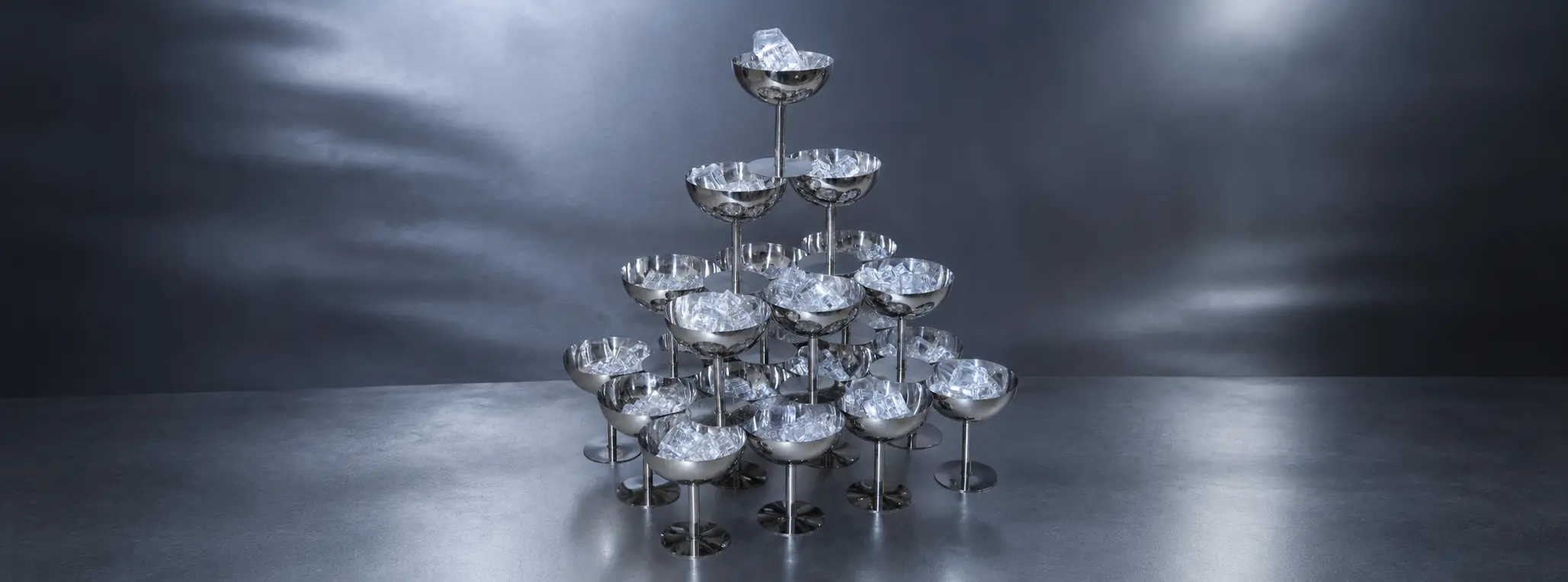 Sculptural champagne tower made of metallic silver coupe glasses arranged in a pyramid against a dramatic monochromatic backdrop.