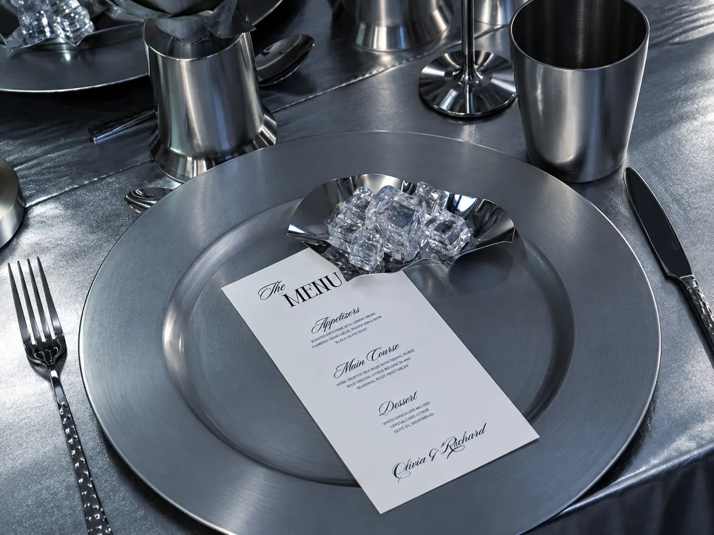 Close-up of a monochromatic silver table setting with a personalized menu, metallic dinnerware, silver cutlery and decorative clear acrylic cubes.