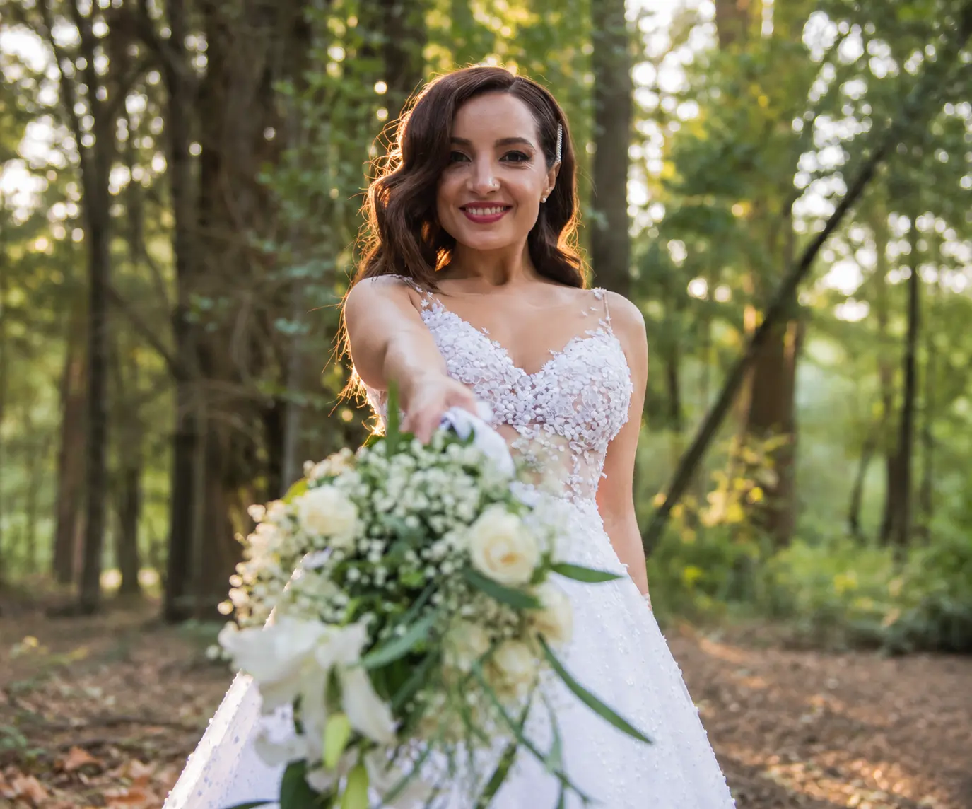 Bride Anna smiling and extending her white floral bouquet toward the camera, wearing a lace wedding gown in a sunlit woodland setting.