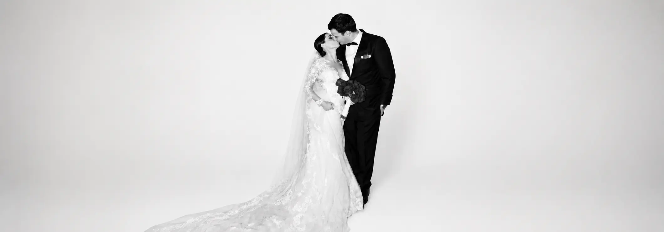 Black and white wedding portrait of a bride and groom kissing, with the bride wearing a lace wedding gown and long flowing train against a minimalist white background.
