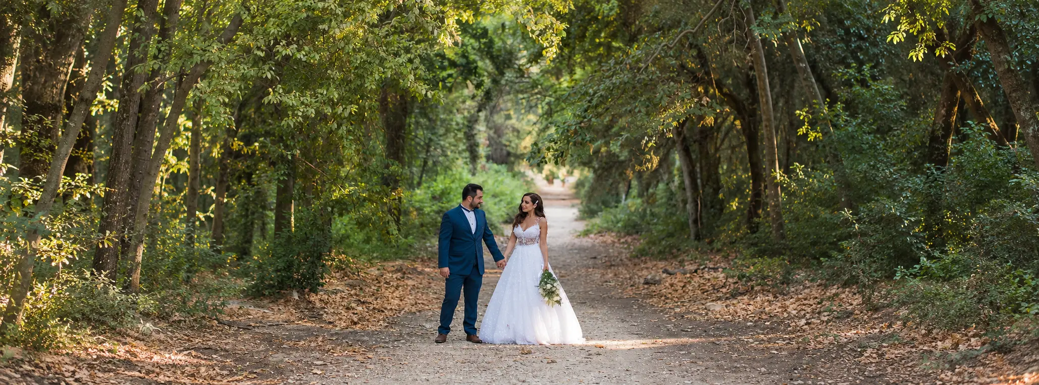 Bride and groom holding hands on a tree-lined woodland path, with the bride wearing a white wedding gown and the groom dressed in a blue suit.