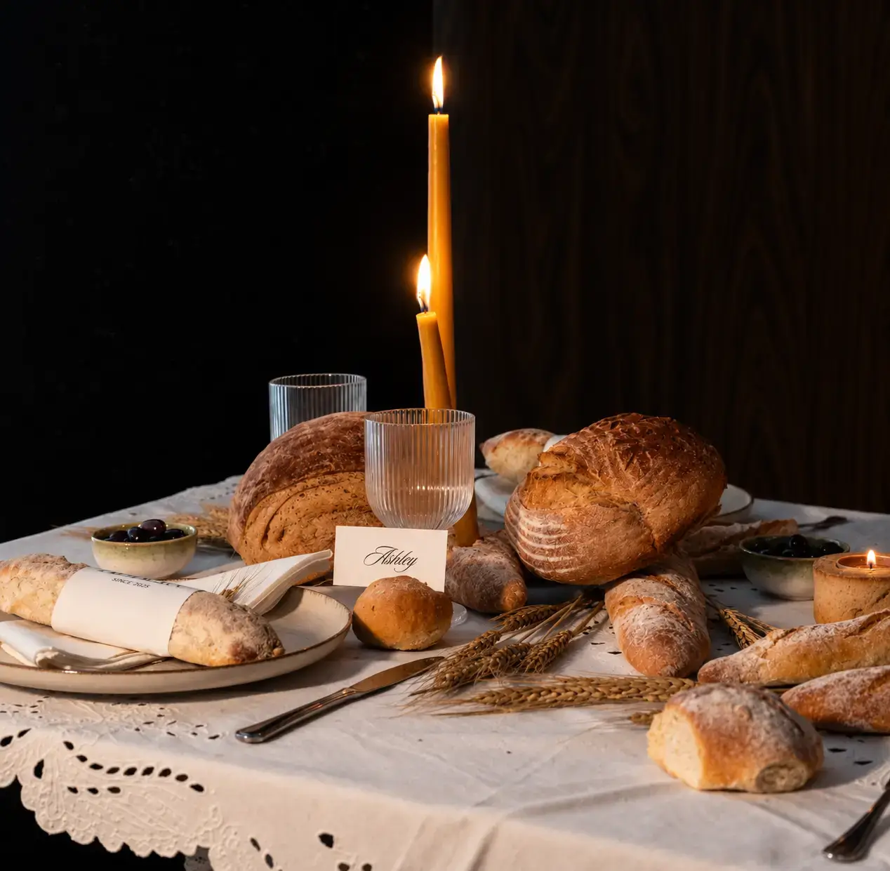 Rustic LEVEIN bread tablescape featuring artisan loaves, baguettes, wheat stalks, vintage glassware and lit taper candles.