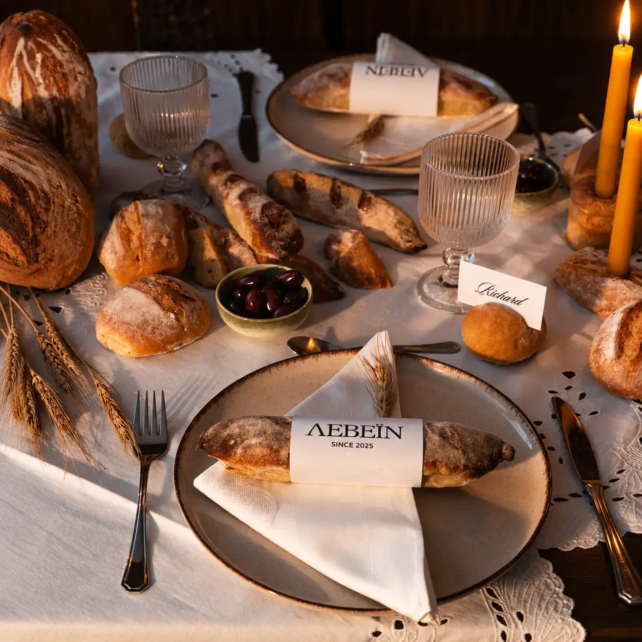LEVEIN artisan bread served on ceramic plates with branded paper bands, surrounded by rustic loaves, olives, wheat stalks and candlelight.