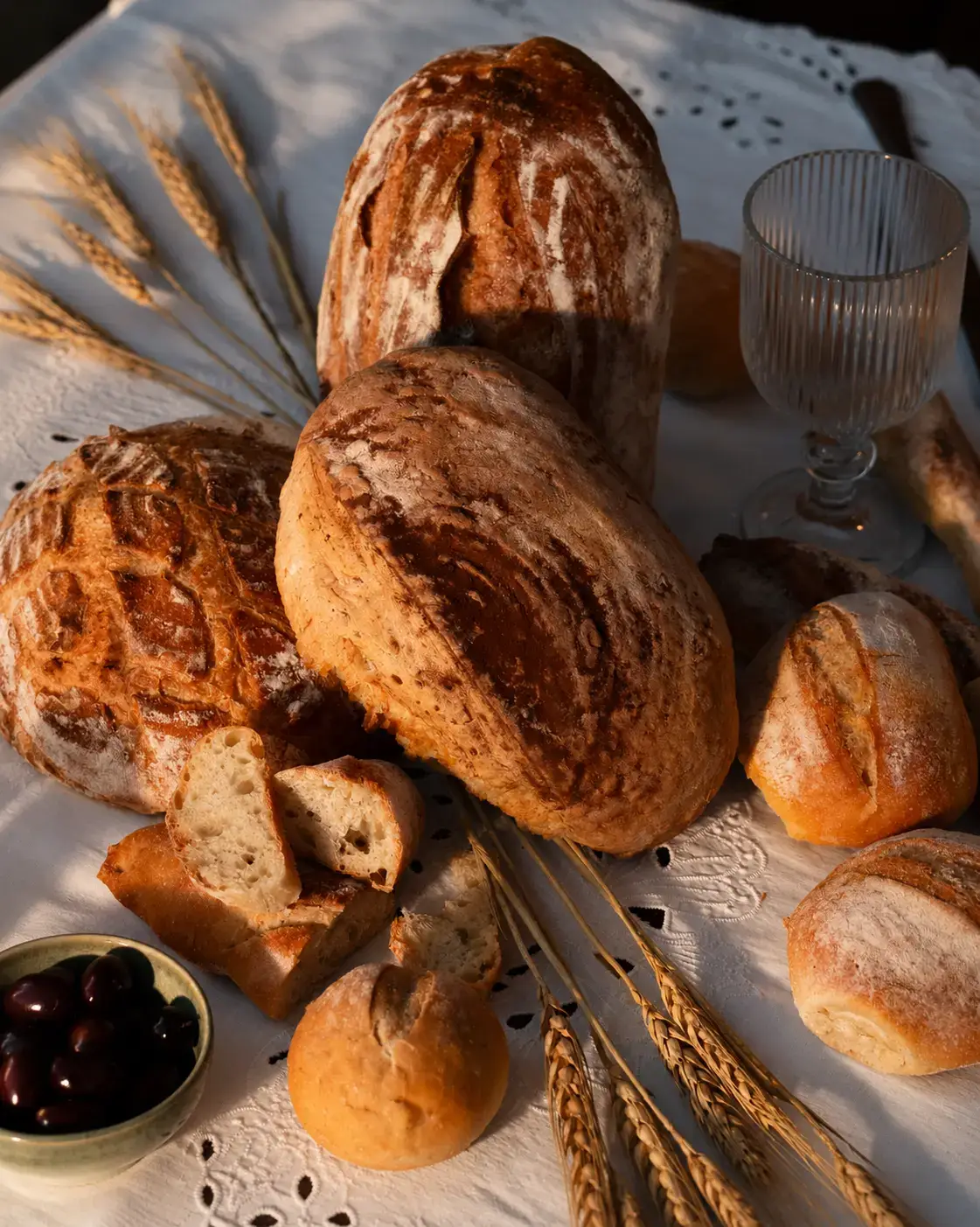 Close-up of rustic artisan bread loaves and sliced sourdough, styled with wheat stalks, olives and vintage glassware.