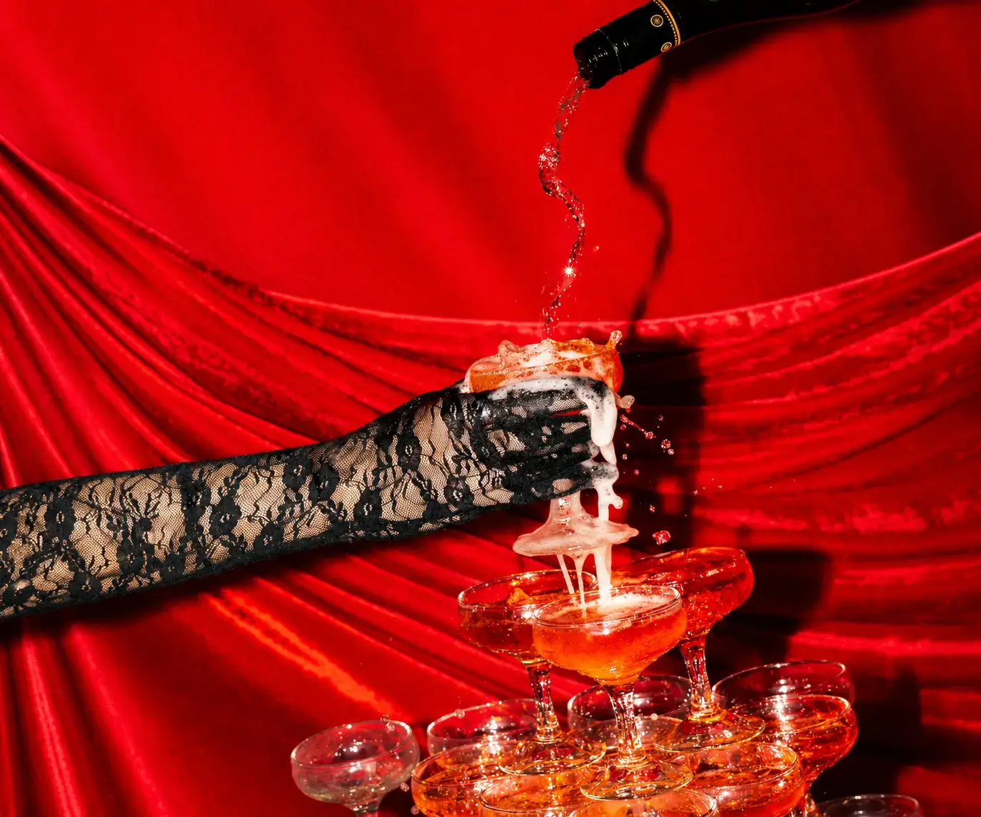 Champagne being poured into a coupe glass held by a black lace-gloved hand above a champagne tower, against a dramatic red satin backdrop.