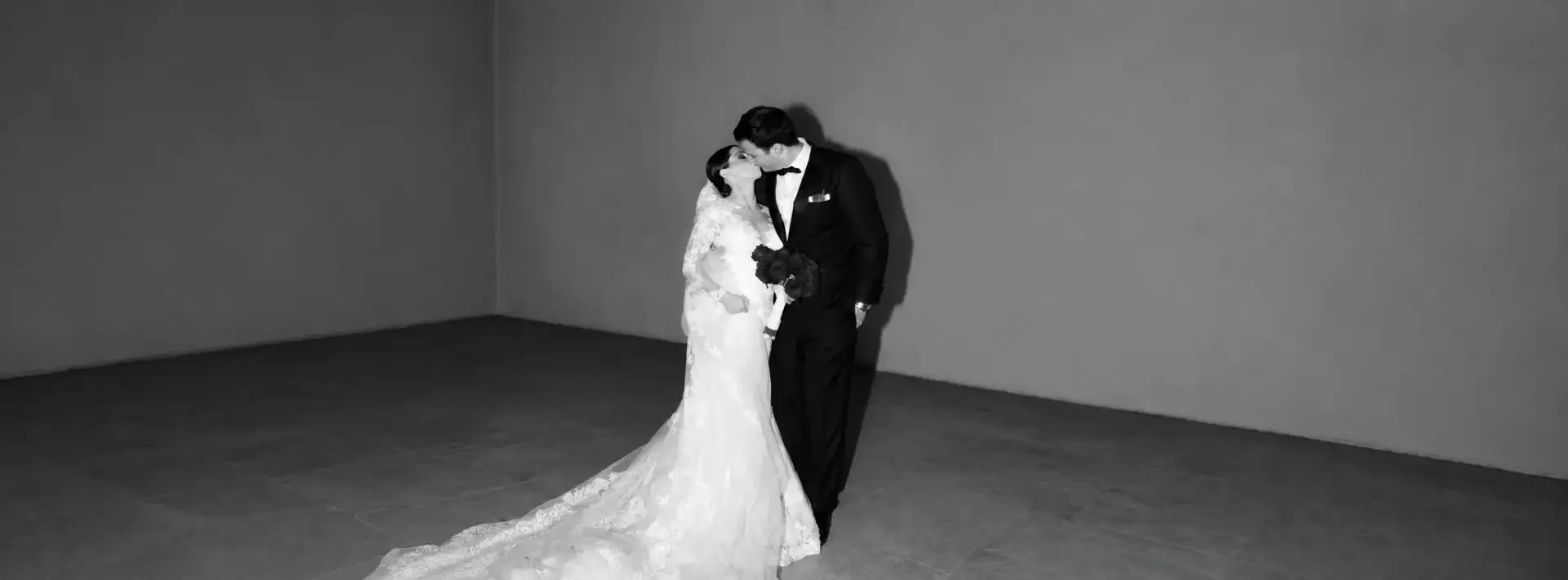 Black and white wedding portrait of the couple sharing a kiss, with the bride wearing a lace gown with a long train and the groom in a black tuxedo.