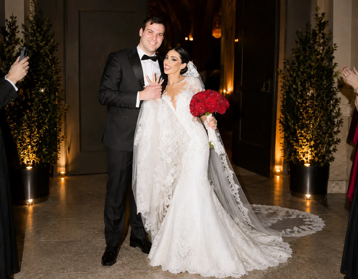 Smiling bride and groom posing together at their wedding reception, with the bride wearing a lace gown and holding a bouquet of red roses.