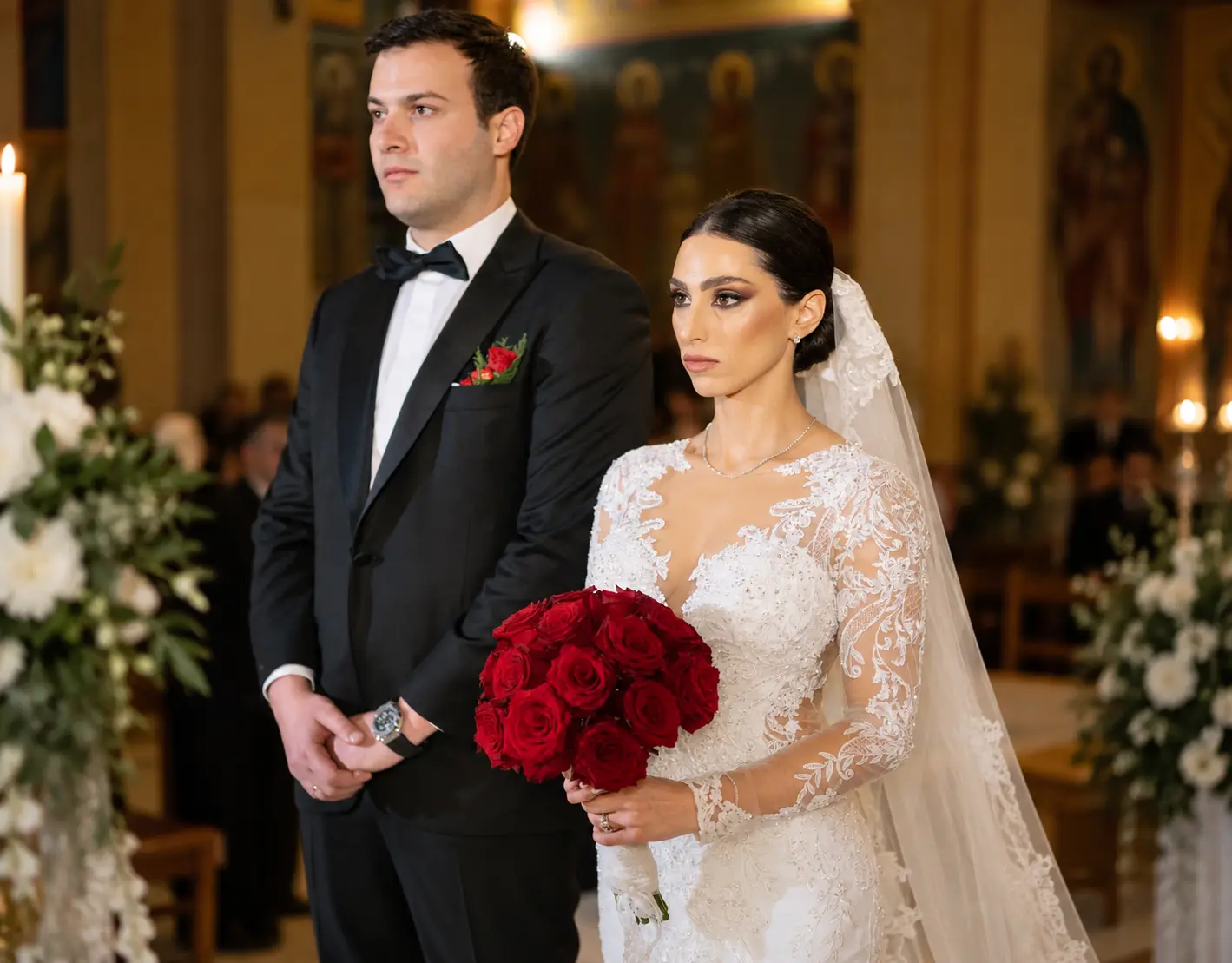 Bride and groom standing together during an Orthodox church wedding ceremony, with the bride wearing a lace gown and holding a bouquet of red roses.