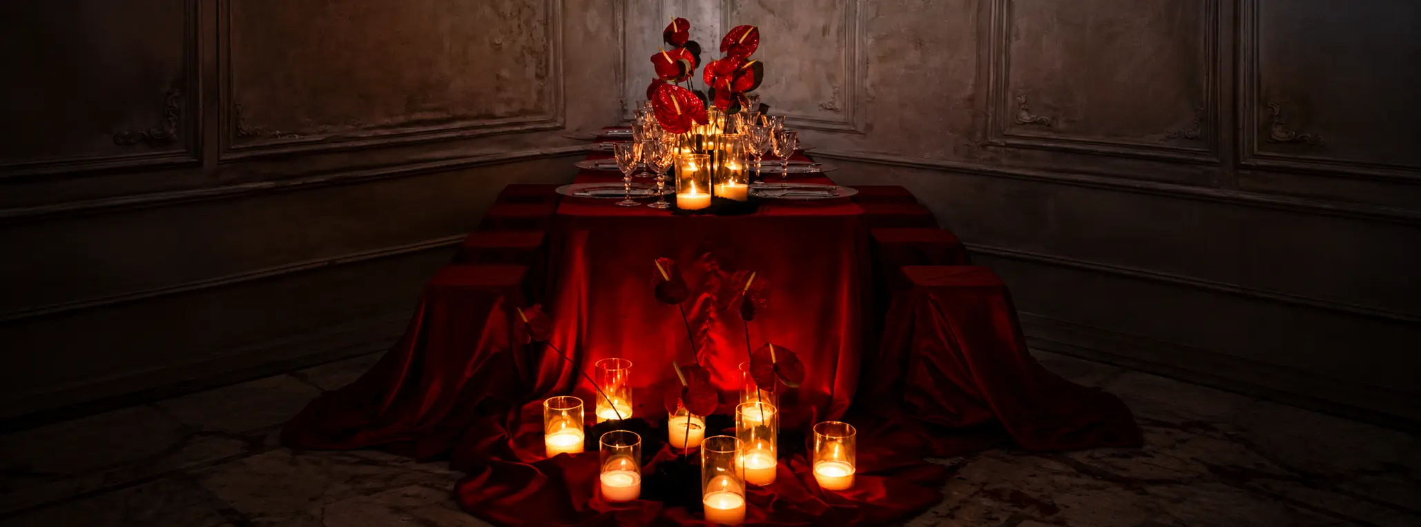 Symmetrical editorial dining table draped in deep red satin, decorated with red anthurium flowers, crystal glassware and glowing candles.