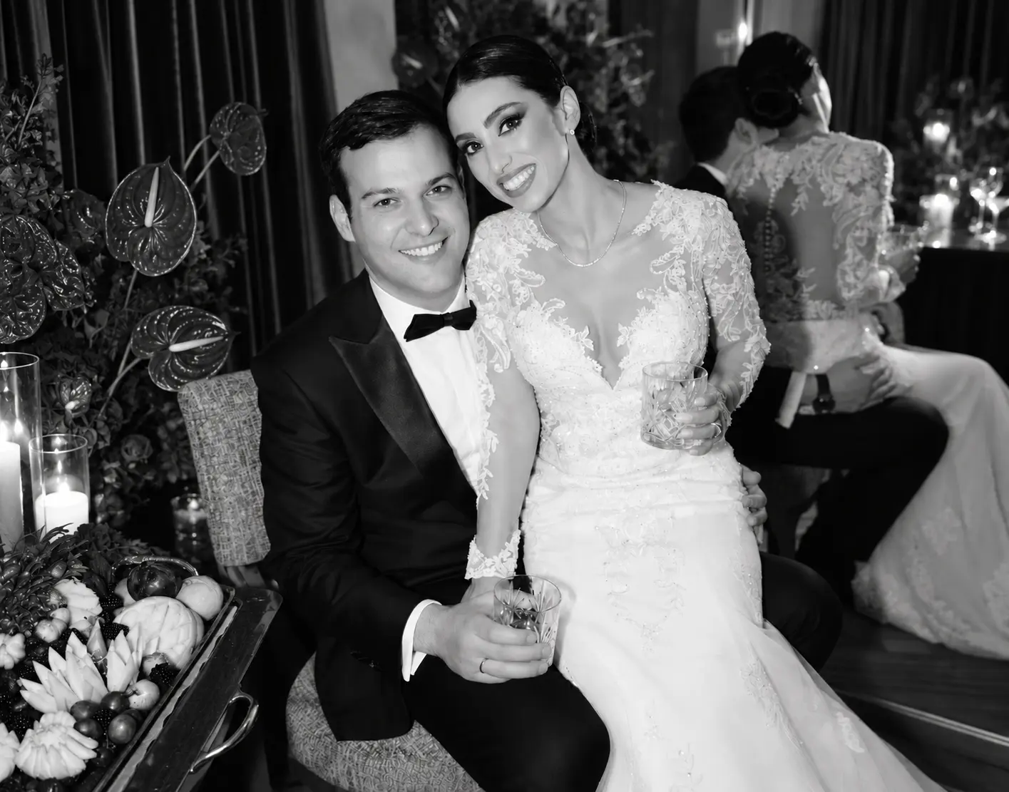 Black and white wedding portrait of a smiling bride and groom seated together at their reception, holding drinks in an elegant setting.
