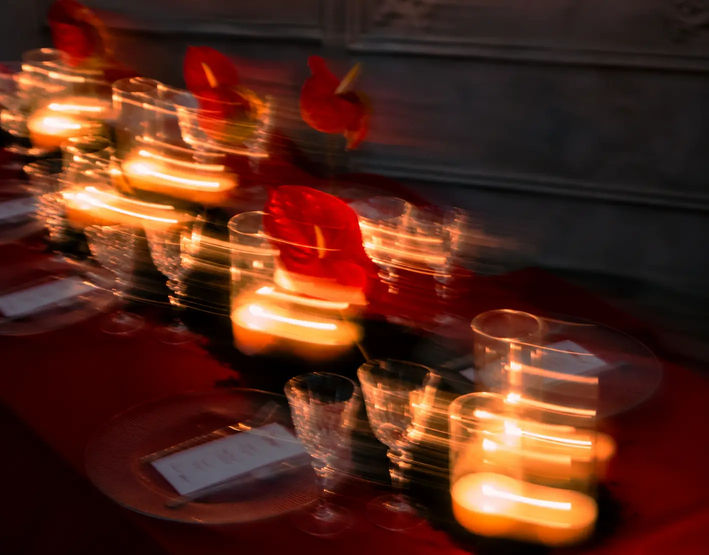 Artistic motion-blur photograph of a red editorial dining table with glowing candles, crystal glassware and red anthurium flowers.