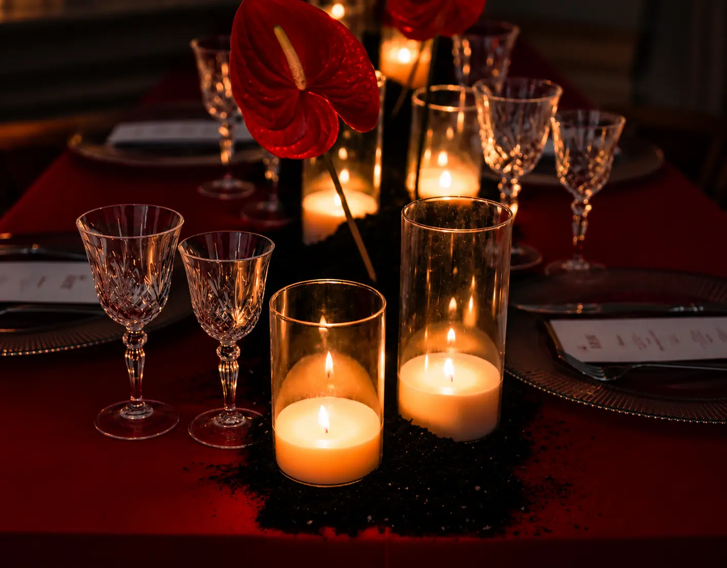 Close-up of glowing candles in glass cylinders, crystal wine glasses and a red anthurium flower on a deep red editorial dining table.