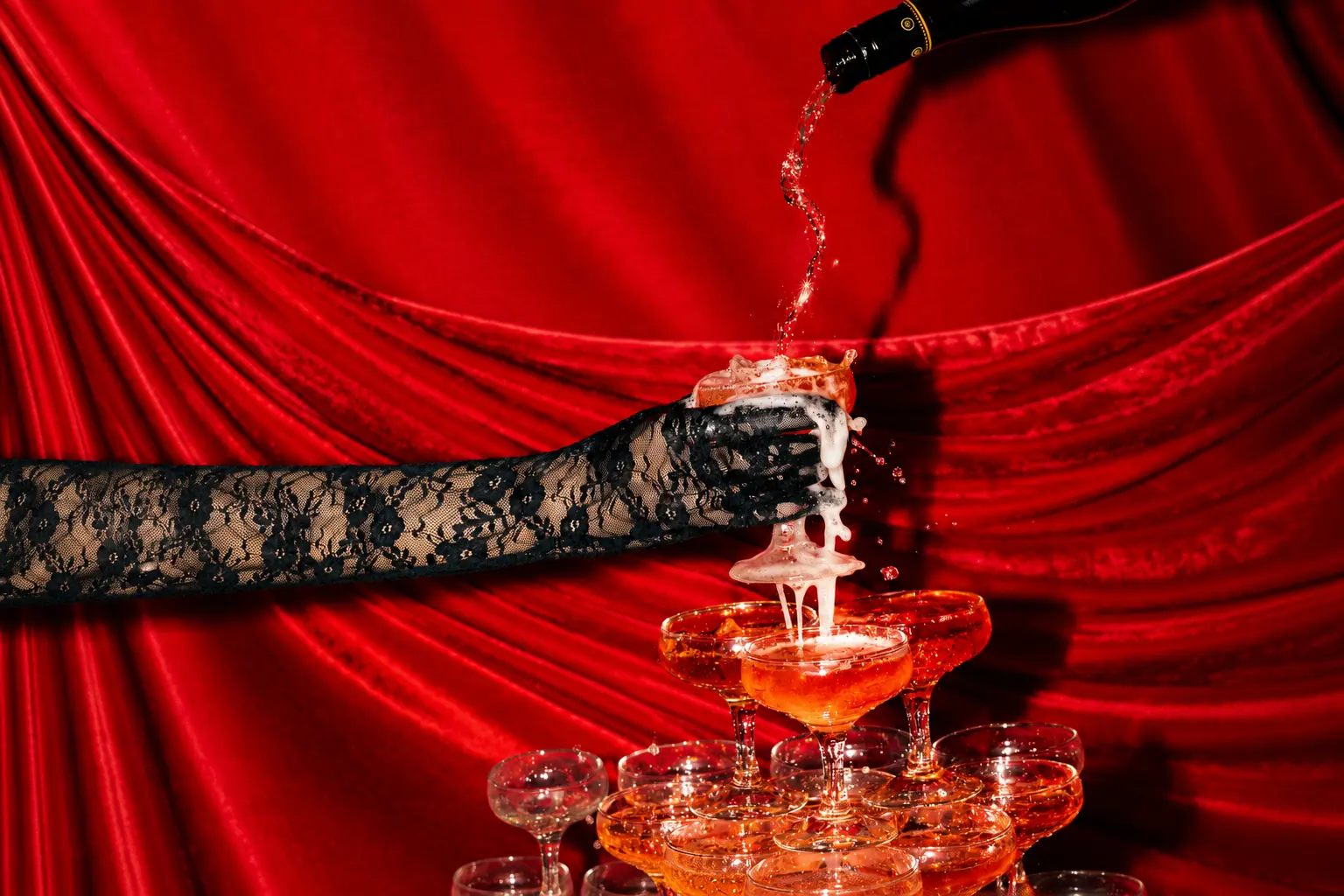 Champagne being poured into a coupe glass held by a black lace-gloved hand above a champagne tower, against rich red satin draping.