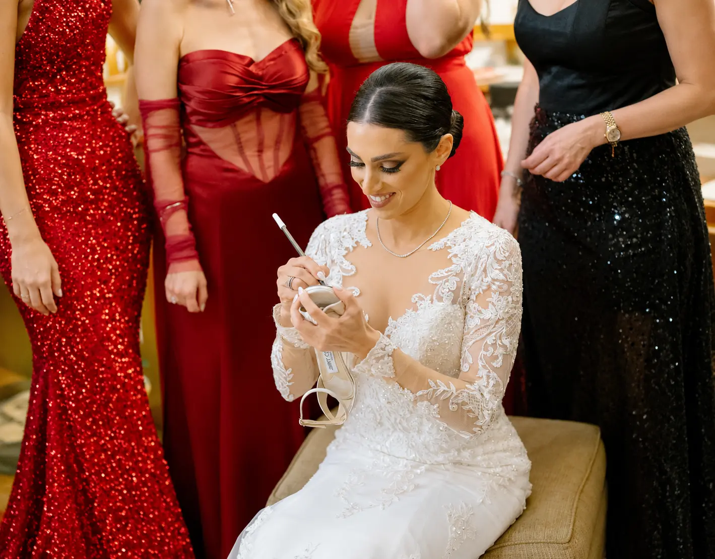Bride in a white lace wedding gown writing on the sole of a bridal shoe, surrounded by women wearing elegant red and black dresses.