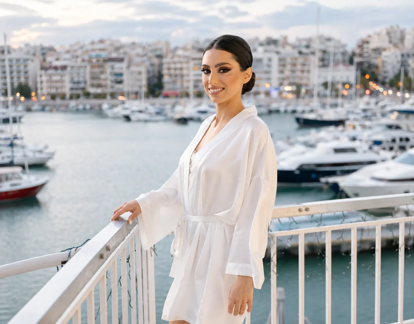 Bride wearing a white satin getting-ready robe on a balcony overlooking a marina filled with yachts and the Athens waterfront.