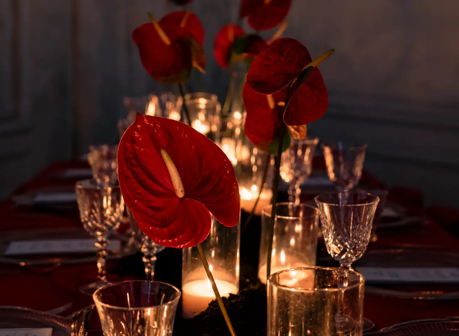 Close-up of red anthurium flowers arranged along an elegant event table with crystal glassware, red linens and glowing candles.