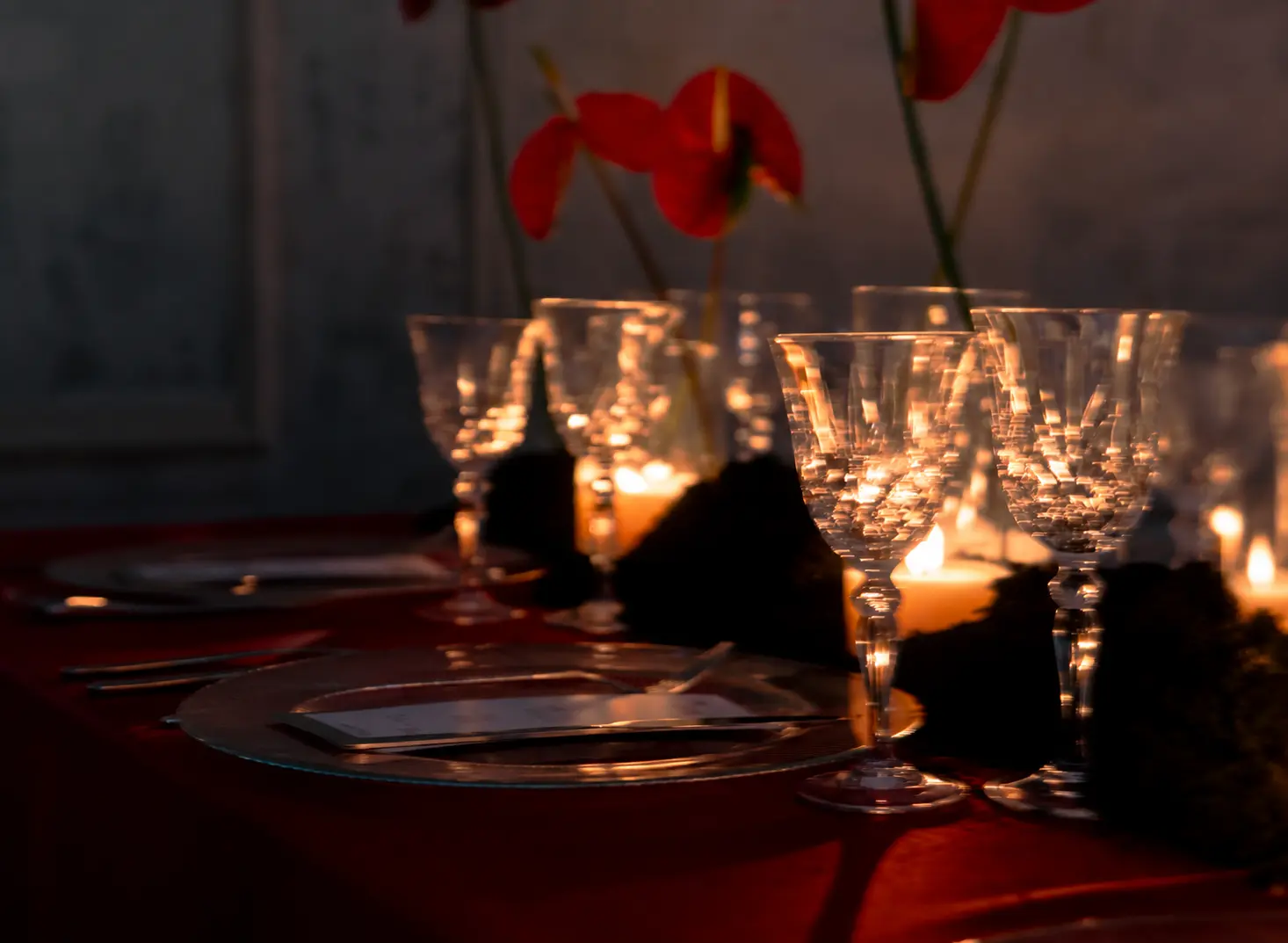 Close-up of crystal wine glasses and candlelit table settings with deep red linens and red anthurium flowers in the background.