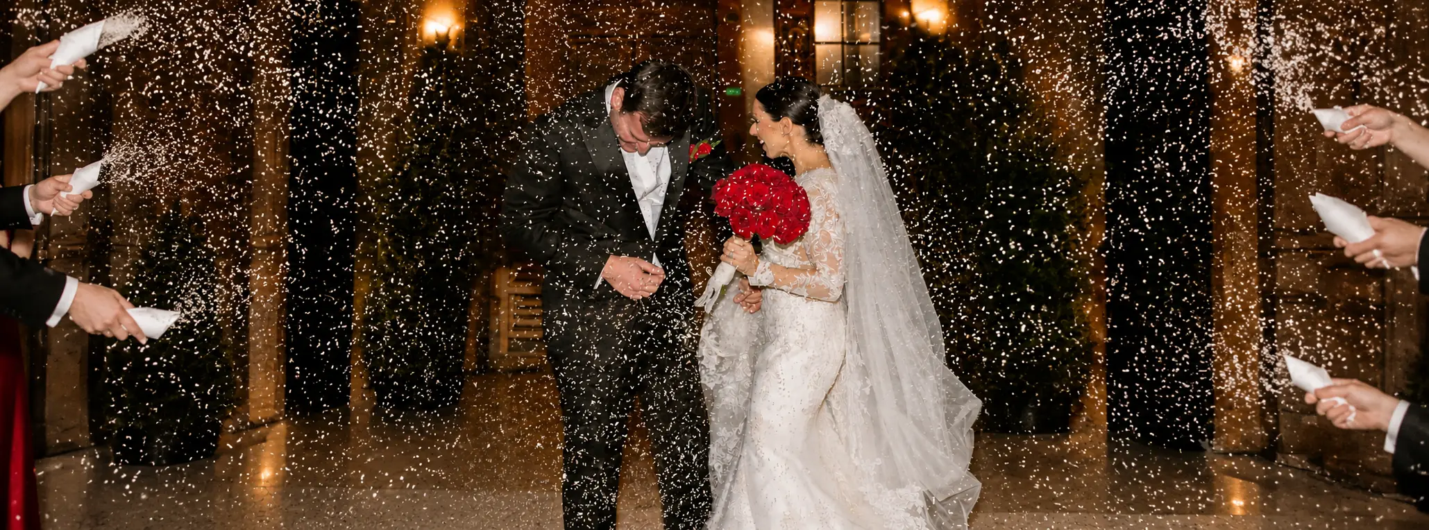 Bride and groom walking through a shower of rice, with the bride holding a bouquet of red roses and wearing a lace wedding gown.