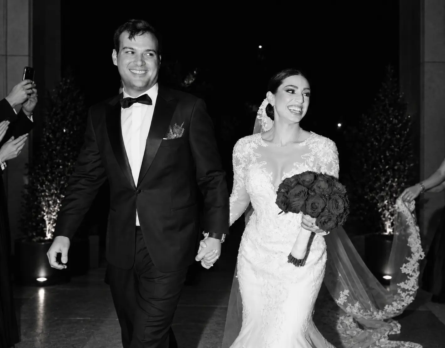 Black and white photograph of a smiling bride and groom walking hand in hand, with the bride holding a rose bouquet and wearing a lace wedding gown.
