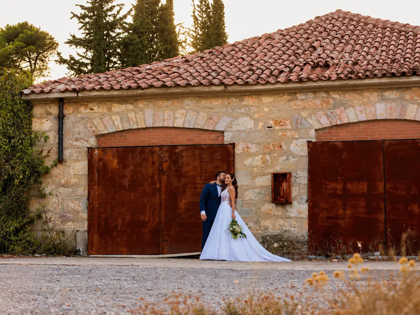 Bride and groom sharing a romantic moment in front of a rustic stone building with terracotta roof tiles and weathered wooden doors.