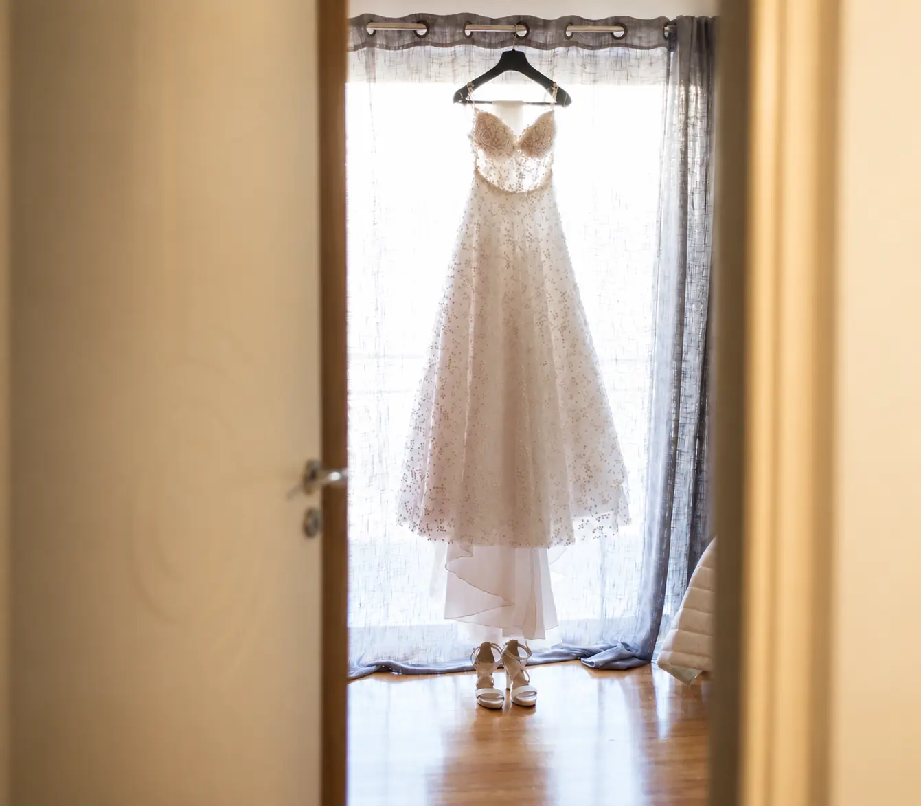 White lace wedding dress hanging in front of a softly lit window, with bridal shoes placed beneath it.