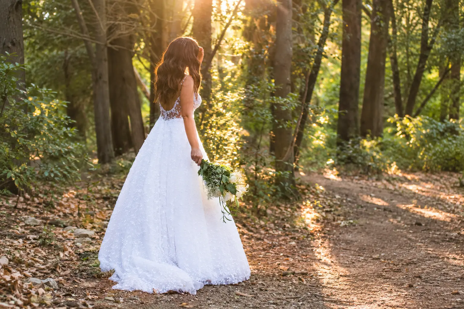 Bride Anna wearing a white lace wedding gown and holding a floral bouquet in a sunlit woodland setting.