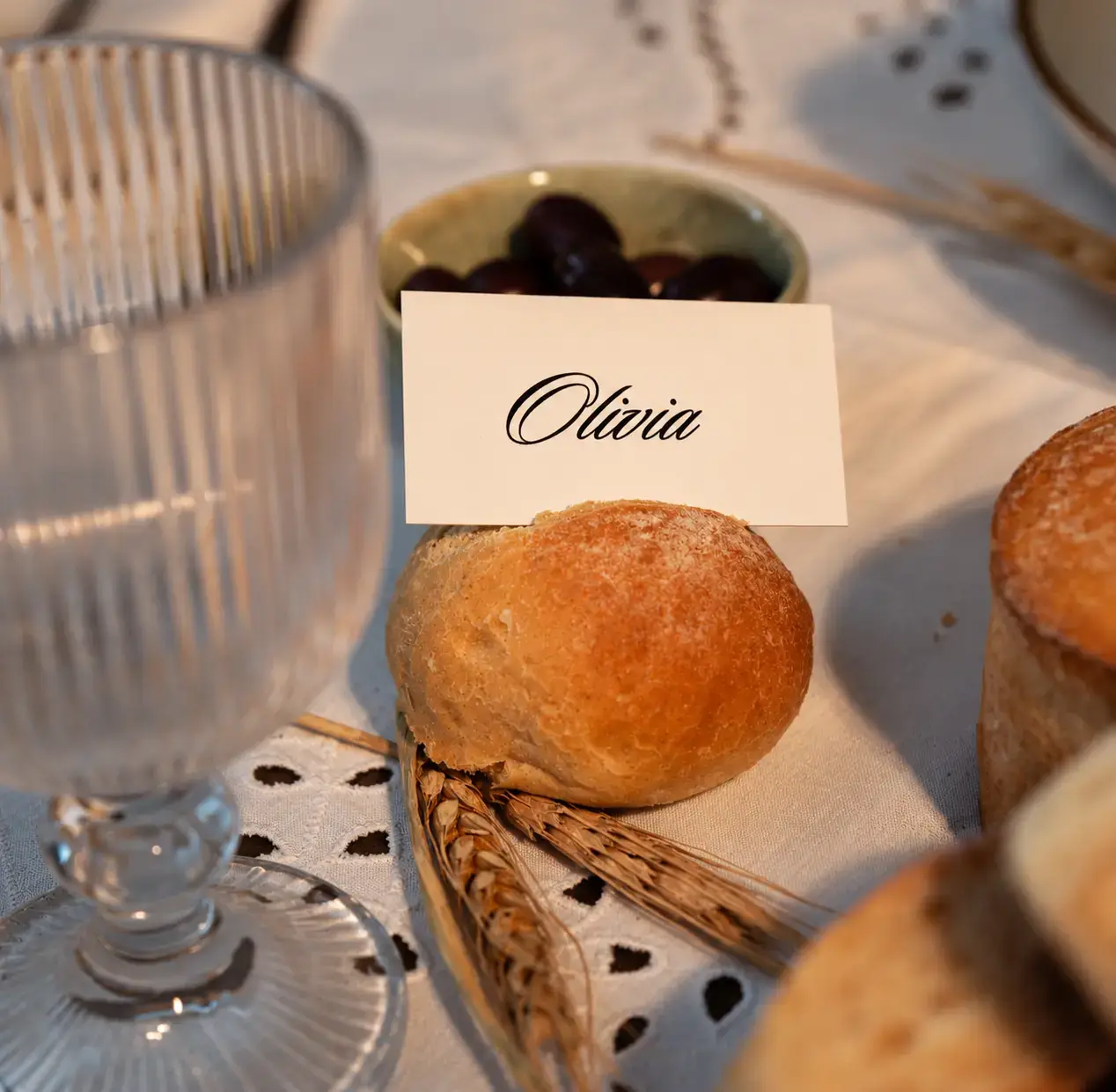 Close-up of an artisan bread roll with a personalized Olivia place card, textured glassware, olives and wheat details at the LEVEIN event