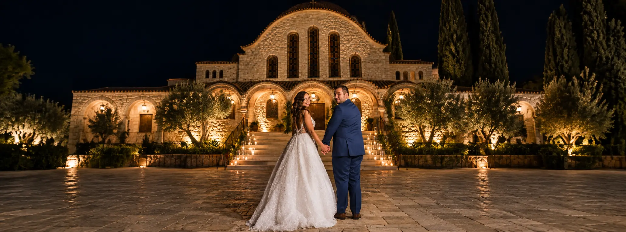 Anna and Antonis holding hands in front of an illuminated stone wedding venue at night, surrounded by candlelit steps and Mediterranean trees.
