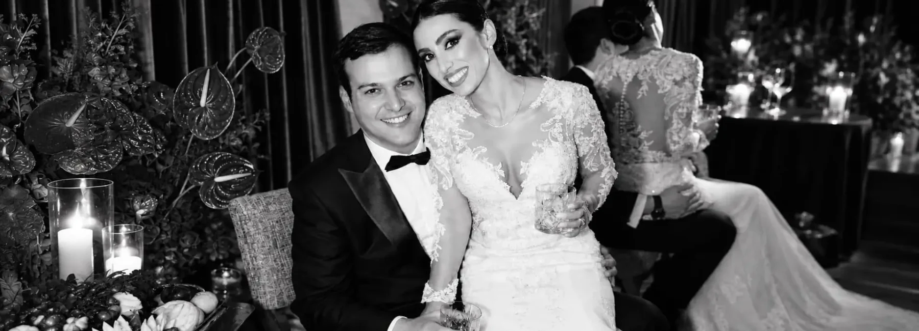 Black and white portrait of a bride and groom smiling together at an elegant wedding reception with floral arrangements and candlelight.